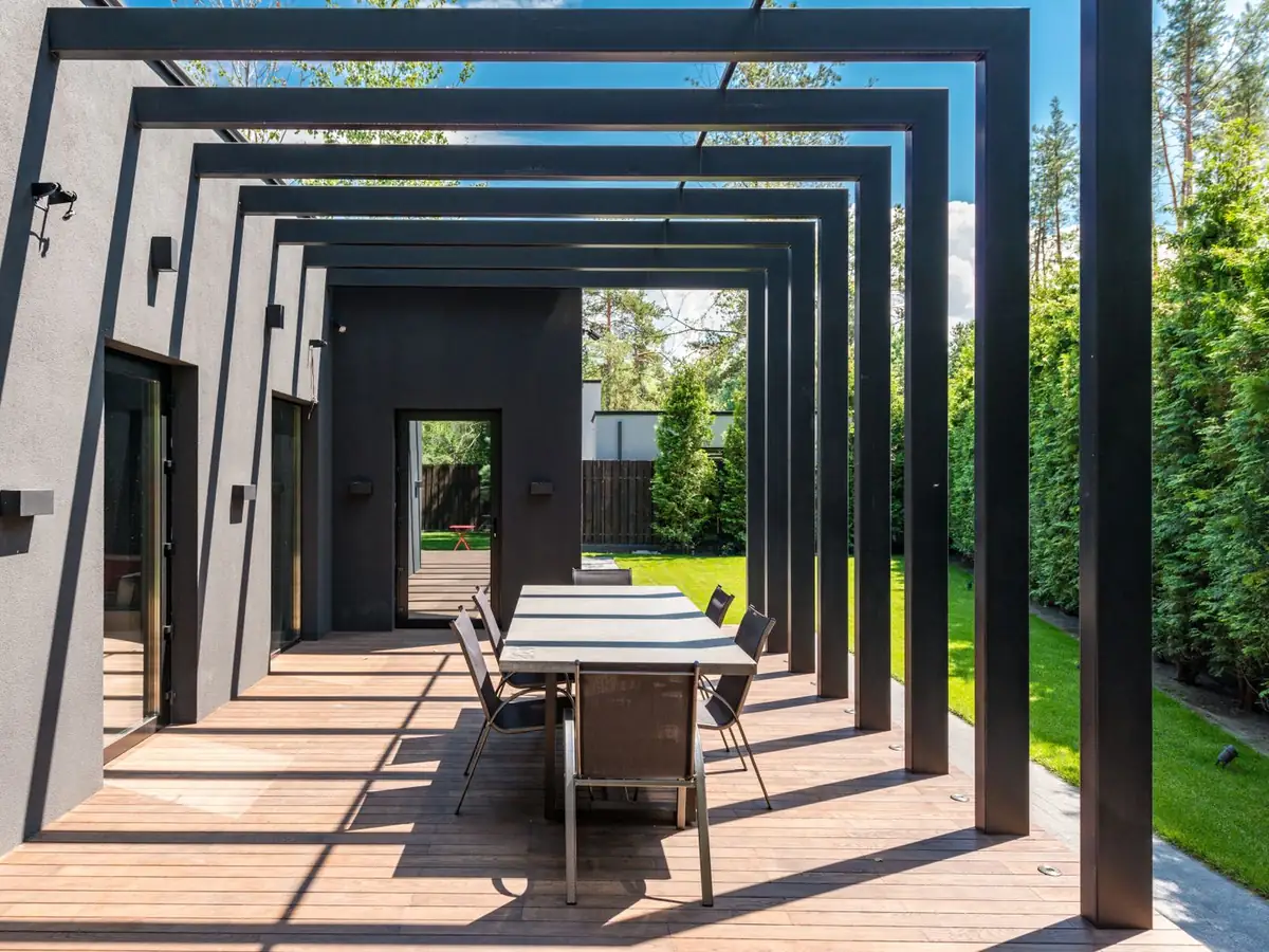 Pérgola de perfiles de acero pintados en negro sobre un deck de madera con mesa y sillas, en el patio de una casa.